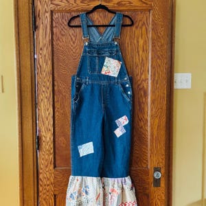 May include: Blue denim overalls with patchwork details. The overalls have a bib pocket, side pockets, and a flared bottom with a patchwork ruffle. The overalls are hanging on a black hanger against a wooden door.