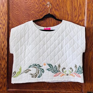 May include: Cream-colored quilted top with a crew neck and short sleeves. The bottom edge features a detailed floral embroidery in shades of green, brown, orange, and pink. The top is hanging on a black hanger.