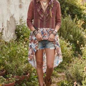 May include: A long-sleeved button-down shirt with a brown plaid bodice, lace sleeves, and a high-low hemline featuring a blue, brown, and white floral print. The outfit includes distressed denim shorts, a wide-brimmed hat, and brown fringe boots.