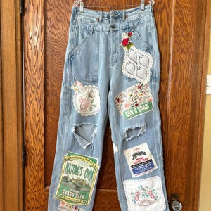 May include: Light blue denim jeans with decorative patches, including floral embroidery, lace, and seed packet labels. The jeans have distressed details and are hanging on a clear hanger.