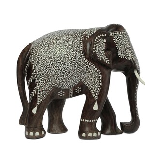 Puede incluir: Una figura de elefante de madera marr&oacute;n oscuro con intrincados patrones de incrustaciones florales blancas. El elefante tiene colmillos de color marfil y est&aacute; de pie sobre cuatro patas. El logotipo "craft by soul" est&aacute; en la esquina superior derecha.