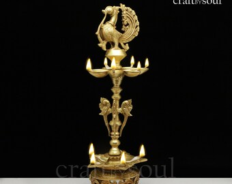May include: A golden brass peacock lamp with multiple oil reservoirs. The lamp has a detailed peacock figure at the top and is supported by an ornate base. The lamp is lit, with flames visible in each reservoir. The base is decorated with orange flower petals.
