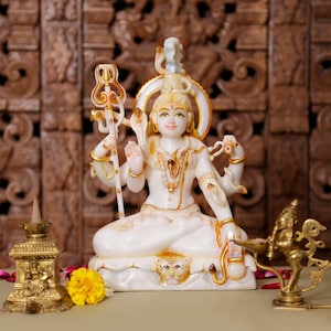 May include: A white and gold statue of a deity with multiple arms, seated in a meditative pose. The statue has intricate details and is adorned with gold accents. A yellow flower and brass objects are in the foreground. The background features a carved wooden panel.