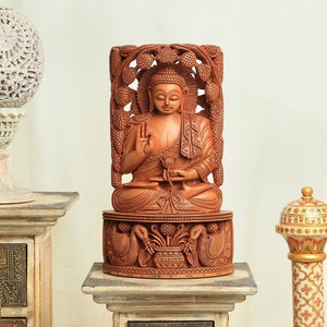 Op de afbeelding: Een gedetailleerd houten Boeddha-beeld, gebeeldhouwd in een zittende positie, met ingewikkelde details. Het beeld heeft een warme bruine kleur en staat op een decoratieve basis met gebeeldhouwde pauwen. De achtergrond is neutraal.