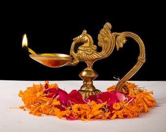 Puede incluir: Una lámpara de aceite de latón con forma de pavo real con una mecha encendida, sobre un fondo negro. La lámpara está rodeada por una guirnalda de pétalos de flores naranjas y rojas. El logotipo de Craft by Soul está en la esquina superior derecha.