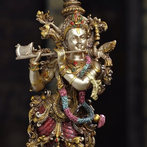 May include: A detailed statue of a deity playing a flute, adorned with intricate gold and red details. The figure stands on a lotus flower base. A small oil lamp with a lit flame is in the foreground.