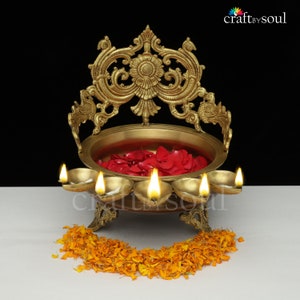 以下が含まれることがあります： 5つの灯芯が灯された真鍮製の装飾的なオイルランプ。中央のボウルには赤いバラの花びらが入り、装飾的な透かし彫りの背面があります。オレンジ色のマリーゴールドの花びらの花輪がベースにあります。 Craft by Soulのロゴ。