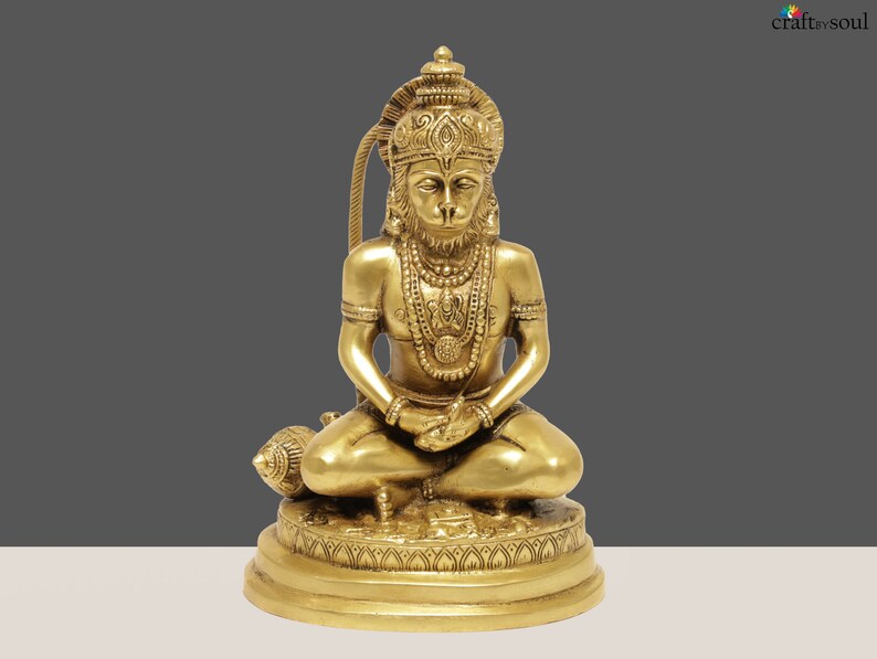 K&ouml;nnte beinhalten: Eine goldene Messingstatue von Hanuman, einer hinduistischen Gottheit, in einer sitzenden Meditationshaltung. Die Statue weist komplizierte Details auf, darunter aufwendiger Schmuck und eine Krone. Die Figur sitzt auf einem detaillierten Sockel, mit einer Keule daneben.