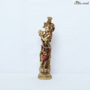 May include: A golden-colored statue of a deity playing a flute. The statue is adorned with intricate details, including a crown, jewelry, and flowing garments in red and gold. The base is a lotus flower design.