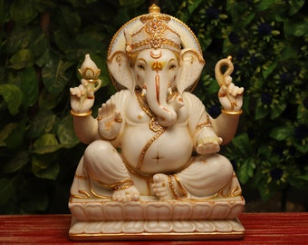 Op de afbeelding: Een gedetailleerd ivoorkleurig beeld van de hindoeïstische godheid Ganesha, zittend op een lotusbasis. Het beeld heeft gouden accenten, ingewikkelde details en een halo. Ganesha houdt een lotusbloem en een lusvormig object in zijn handen.