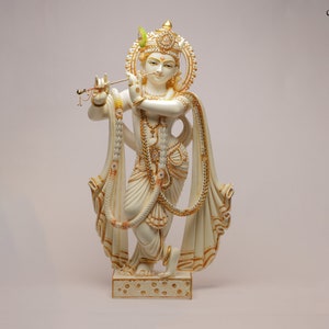 Puede incluir: Una estatua crema de una deidad, adornada con detalles dorados e intrincados. La figura sostiene una flauta y lleva joyas ornamentadas y un tocado decorativo. La estatua se encuentra sobre una base detallada, con el texto "craft by soul" visible.