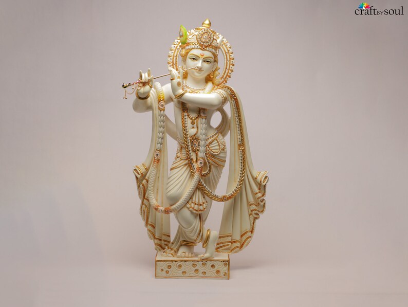 Puede incluir: Una estatua crema de una deidad, adornada con detalles dorados e intrincados. La figura sostiene una flauta y lleva joyas ornamentadas y un tocado decorativo. La estatua se encuentra sobre una base detallada, con el texto "craft by soul" visible.