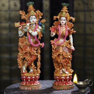 May include: Two ornate statues of Hindu deities. The figures are adorned with gold and red detailing, white skin, and pink garlands. One holds a flute and the other a hand gesture. They stand on lotus flower bases, with a small oil lamp in the foreground.
