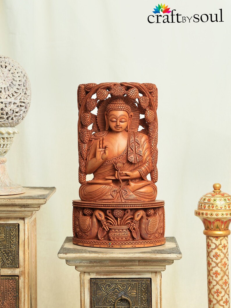 Op de afbeelding: Een gedetailleerd houten Boeddha-beeld, gebeeldhouwd in een zittende positie, met ingewikkelde details. Het beeld heeft een warme bruine kleur en staat op een decoratieve basis met gebeeldhouwde pauwen. De achtergrond is neutraal.