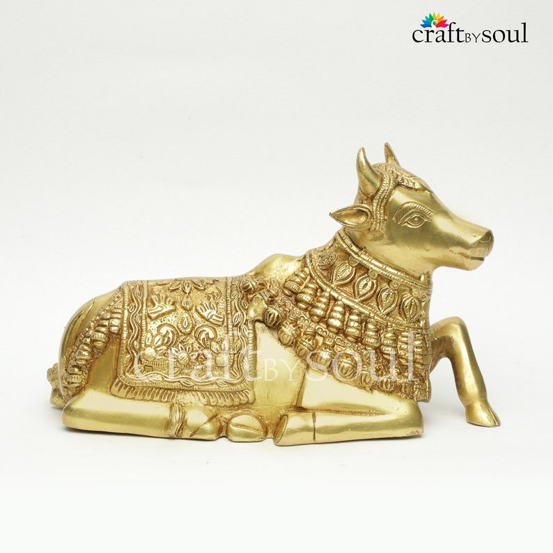 May include: A golden-colored brass sculpture of a reclining cow, adorned with intricate engraved patterns on its body and head. The cow is in a resting position, with its legs tucked beneath. The sculpture has a detailed design and is set against a white background.