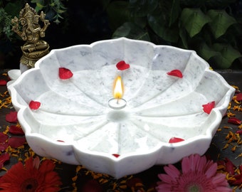 Puede incluir: Un cuenco de mármol blanco con forma de loto con una vela encendida en el centro. Pétalos de rosa rojos están esparcidos dentro del cuenco. El cuenco está rodeado de flores rojas y rosas y una pequeña estatua dorada.