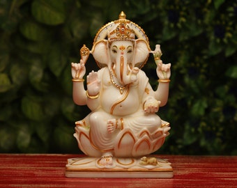 May include: A white and gold statue of the Hindu deity Ganesha, seated on a lotus flower. The statue features intricate details, including a crown, jewelry, and a small mouse at the base. The background is a blurred green foliage.