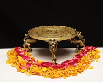 May include: A brass decorative stand with a round top and ornate detailing. The stand is supported by three sculpted animal figures. The stand is surrounded by a garland of orange and red flower petals, set against a black background.
