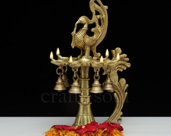May include: A brass peacock-shaped oil lamp with seven candle holders. The lamp features a detailed peacock design, hanging bells, and is adorned with red rose petals and orange marigold flowers. The Craft by Soul logo is visible in the top right corner.