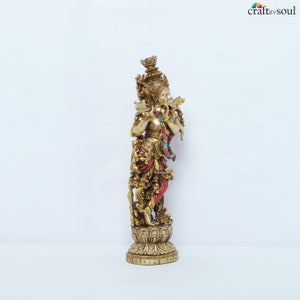 May include: A gold-colored statue of a deity playing a flute. The statue has intricate details, including a crown, ornate clothing with red and gold accents, and a lotus flower base. The statue is set against a white background.