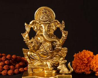 May include: A golden brass statue of the Hindu deity Ganesha, seated with a rat at his feet. The statue has intricate details and is set against a dark background, with orange flowers and a string of beads on either side.