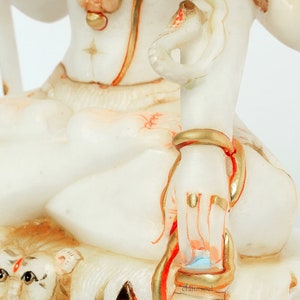 May include: A white statue of a deity with gold and red accents. The statue features intricate details, including a snake wrapped around an arm and a lion's head at the base. The statue has a serene expression and is adorned with jewelry.