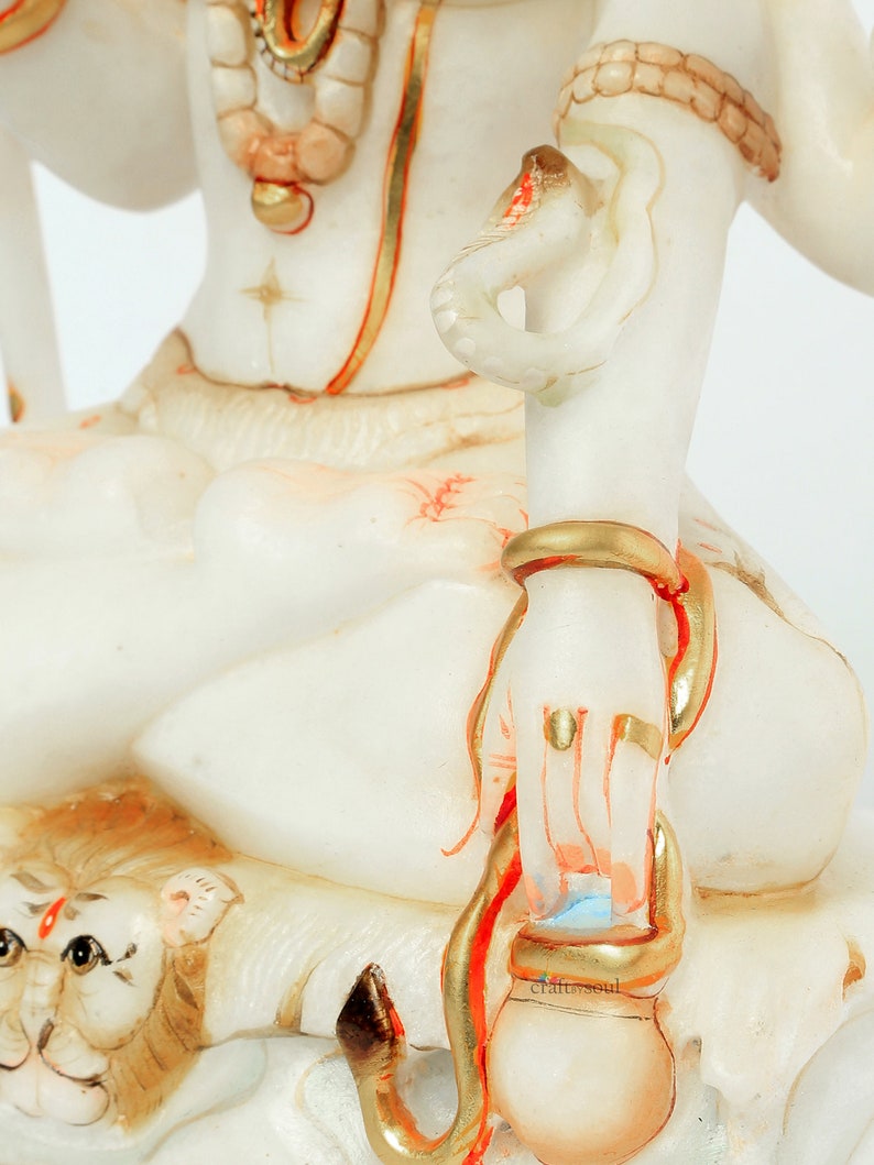 May include: A white statue of a deity with gold and red accents. The statue features intricate details, including a snake wrapped around an arm and a lion's head at the base. The statue has a serene expression and is adorned with jewelry.