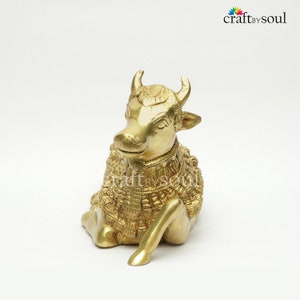 May include: A golden brass sculpture of a seated cow with detailed ornamentation. The cow has curved horns and is in a resting position. The sculpture is on a white background. The Craft by Soul logo is in the upper right corner.