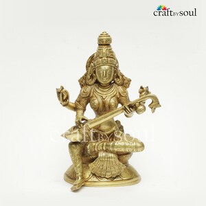 Puede incluir: Estatua de lat&oacute;n de una deidad sentada tocando un instrumento de cuerda. La figura est&aacute; adornada con joyas detalladas y una toca. La estatua tiene un tono dorado y est&aacute; sobre un fondo blanco.
