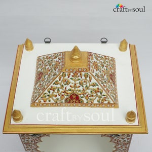 以下が含まれることがあります： 白と金の装飾的な祭壇で、ピラミッド型のトップが付いています。祭壇には、赤、緑、金色の複雑な花の模様が施されています。縁は金でトリミングされ、上部には4つの金の尖塔があります。「craft by soul」というテキストが表示されます。