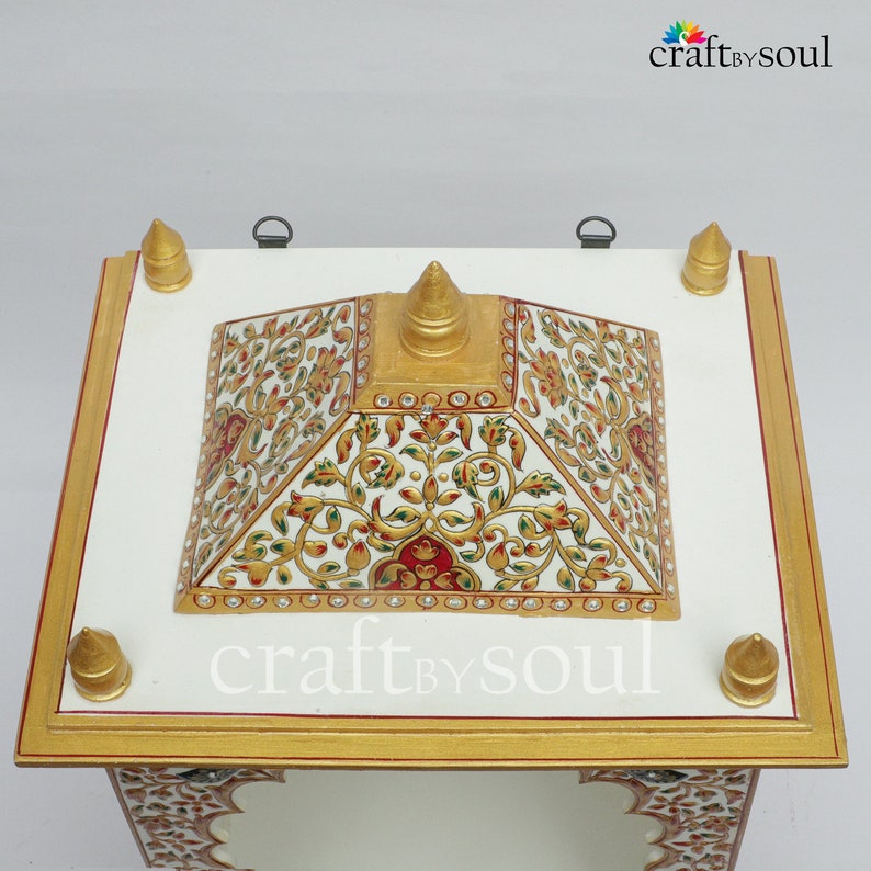 以下が含まれることがあります： 白と金の装飾的な祭壇で、ピラミッド型のトップが付いています。祭壇には、赤、緑、金色の複雑な花の模様が施されています。縁は金でトリミングされ、上部には4つの金の尖塔があります。「craft by soul」というテキストが表示されます。