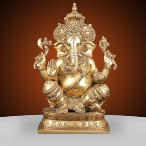 May include: A golden brass statue of the Hindu deity Ganesha, seated in a cross-legged pose. The statue features intricate details, including a crown, multiple arms holding various objects, and a detailed base. The background is a gradient of brown and white.