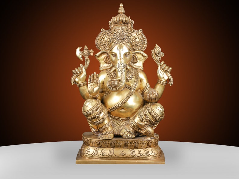 May include: A golden brass statue of the Hindu deity Ganesha, seated in a cross-legged pose. The statue features intricate details, including a crown, multiple arms holding various objects, and a detailed base. The background is a gradient of brown and white.