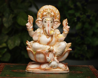 May include: A detailed ivory-colored statue of the Hindu deity Ganesha, seated on a lotus flower. The statue features gold accents, intricate detailing, and a small mouse at the base. The background is a blurred green.