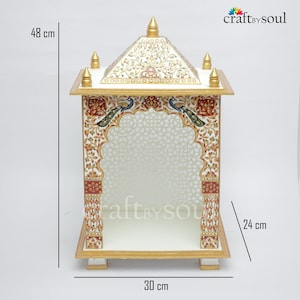以下が含まれることがあります： 高さ48cm、幅30cm、奥行き24cmの白と金の装飾寺院。寺院には、赤、緑、青の複雑な花柄と孔雀のデザインが施されています。屋根には金のアクセントと尖塔があります。内部には模様があります。