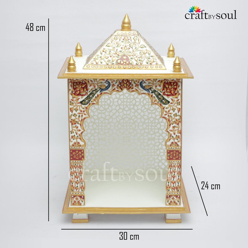 以下が含まれることがあります： 高さ48cm、幅30cm、奥行き24cmの白と金の装飾寺院。寺院には、赤、緑、青の複雑な花柄と孔雀のデザインが施されています。屋根には金のアクセントと尖塔があります。内部には模様があります。
