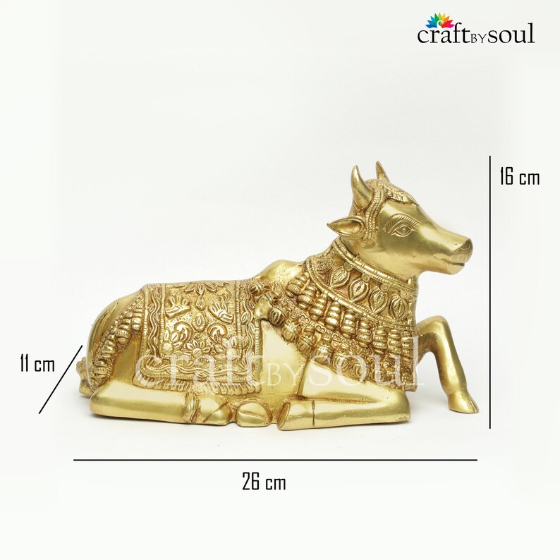 May include: A golden-colored brass cow statue, 26 cm long, 11 cm high, and 16 cm tall. The cow is in a resting position, with detailed carvings on its body and decorative elements around its neck. The product is from craftbysoul.