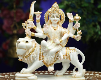 May include: A white and gold statue depicting a multi-armed deity on a lion. The statue features detailed gold embellishments on the lion's mane and the deity's adornments. The deity is seated, with multiple arms holding various items.