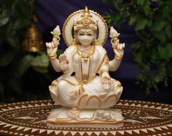 May include: A white and gold statue of a seated deity, adorned with a halo and intricate jewelry. The figure is seated on a lotus flower, with a small animal at its base. The statue has a serene expression and is set against a blurred background.