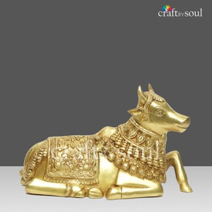 May include: A golden brass sculpture of a reclining cow, adorned with intricate floral and geometric patterns. The cow is wearing decorative jewelry and a blanket. The sculpture is a detailed representation of a sacred animal.