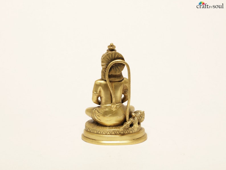 K&ouml;nnte beinhalten: Eine goldfarbene Messingstatue einer sitzenden Figur, von hinten gesehen. Die Figur hat einen aufwendigen Kopfschmuck und sitzt auf einem detaillierten kreisf&ouml;rmigen Sockel. Ein gebogenes, dekoratives Element erstreckt sich vom R&uuml;cken der Figur. Wei&szlig;er Hintergrund.