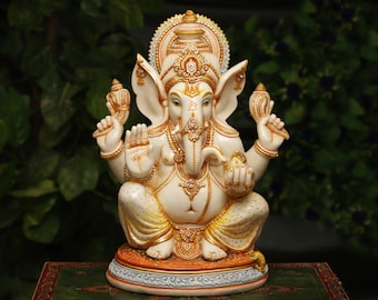 May include: A detailed ivory-colored statue of the Hindu deity Ganesha, seated with multiple arms and an elephant head. The statue features gold and jewel accents, intricate detailing, and a halo. The background is a blurred green.