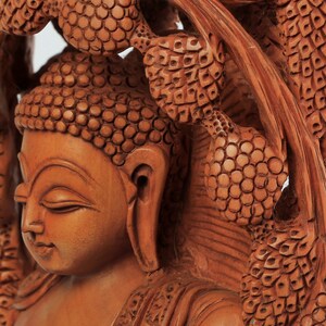 Op de afbeelding: Een gedetailleerde houten sculptuur van Boeddha met gesloten ogen, gesneden in een warme bruine tint. De sculptuur is omgeven door ingewikkeld gesneden bladeren, die de vaardigheid van de kunstenaar laten zien. Het stuk roept een gevoel van vrede en sereniteit op.