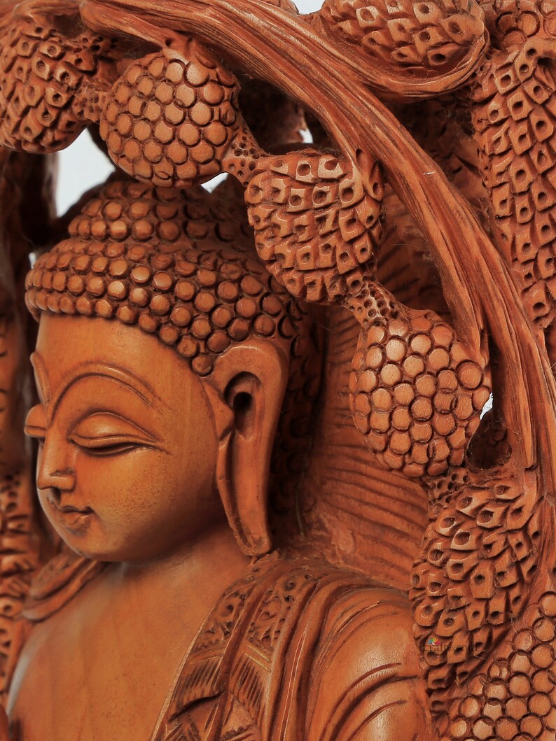 Op de afbeelding: Een gedetailleerde houten sculptuur van Boeddha met gesloten ogen, gesneden in een warme bruine tint. De sculptuur is omgeven door ingewikkeld gesneden bladeren, die de vaardigheid van de kunstenaar laten zien. Het stuk roept een gevoel van vrede en sereniteit op.