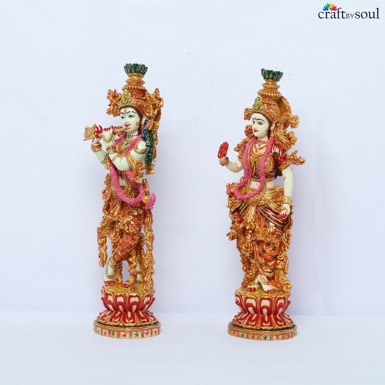 May include: Two ornate statues of Hindu deities. The statues are predominantly gold, red, and cream-colored, with intricate detailing. One figure holds a flute, while the other has a hand raised in a blessing gesture. Both are adorned with pink garlands and stand on decorative bases.