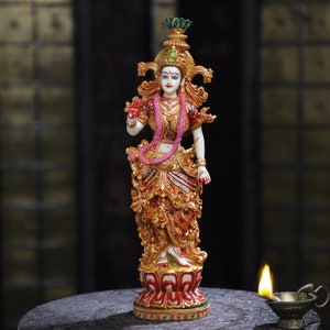 May include: A detailed statue of a deity, featuring a white complexion, adorned with gold and red clothing, and a pink garland. The statue stands on a red and gold lotus base, with intricate details throughout. A small oil lamp is lit in the foreground.