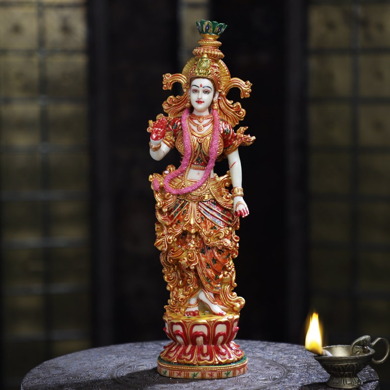 May include: A detailed statue of a deity, featuring a white complexion, adorned with gold and red clothing, and a pink garland. The statue stands on a red and gold lotus base, with intricate details throughout. A small oil lamp is lit in the foreground.