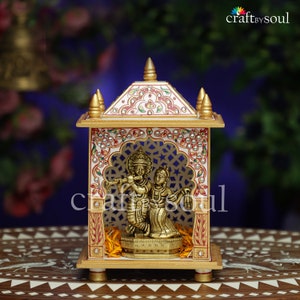 Puede incluir: Un templo decorativo de mármol con detalles dorados y una estatua de latón de deidades hindúes. El templo presenta intrincados patrones en rojo, blanco y dorado, con un diseño de celosía. La base es de madera y el diseño es ornamentado.