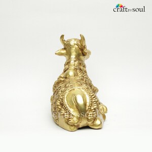 May include: A golden brass cow figurine, viewed from the rear, showcasing intricate embossed detailing. The sculpture features curved horns and ornate neck adornments. The Craft by Soul logo is visible in the upper right corner.