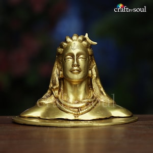 Può includere: Busto dorato di una divinità con occhi chiusi e lunghi capelli fluenti. La scultura presenta dettagli intricati, tra cui un simbolo del terzo occhio sulla fronte e una collana. Il busto è posto su una base rotonda.